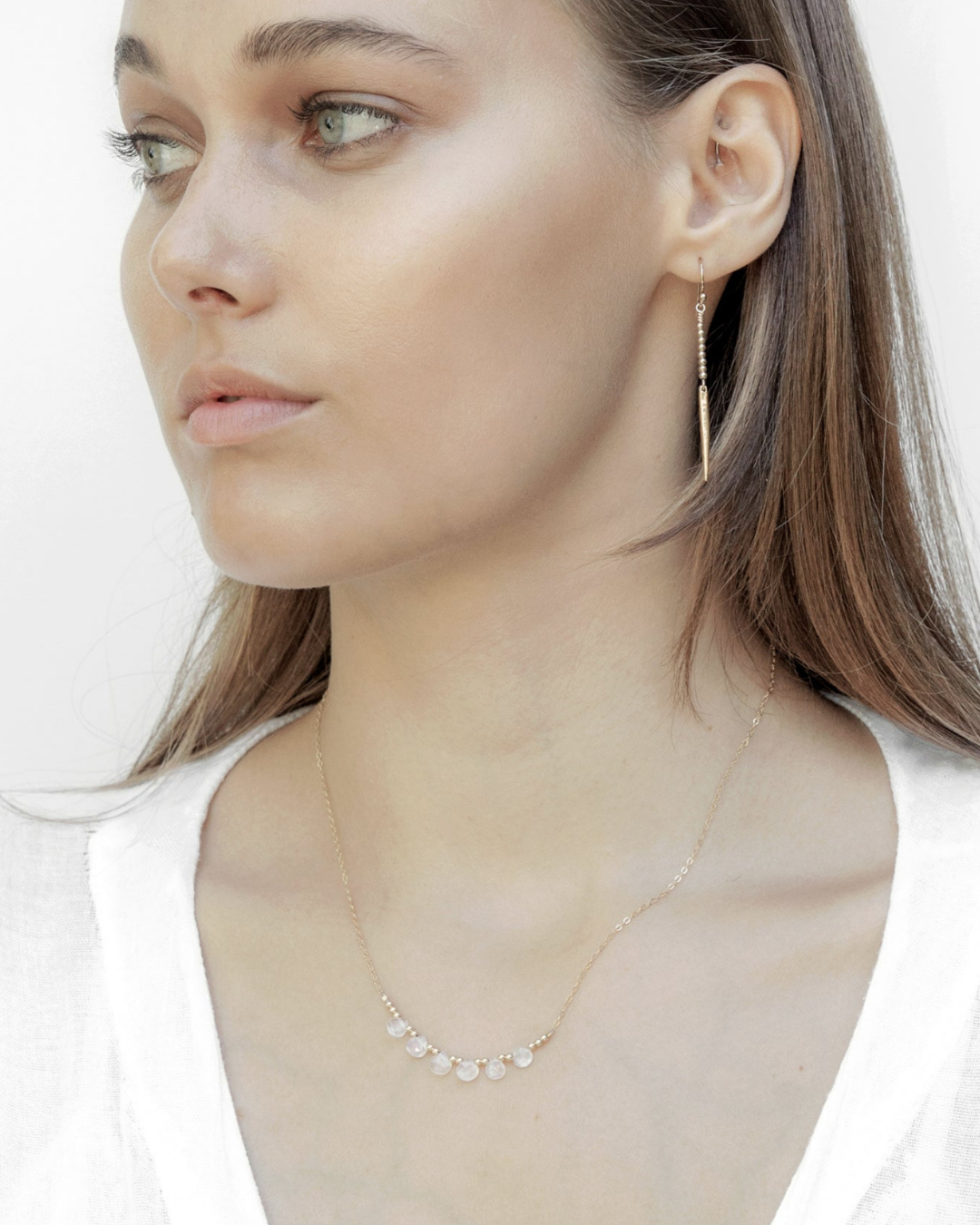 Close-up of a woman wearing gold earrings and a delicate necklace with moonstones on a white background