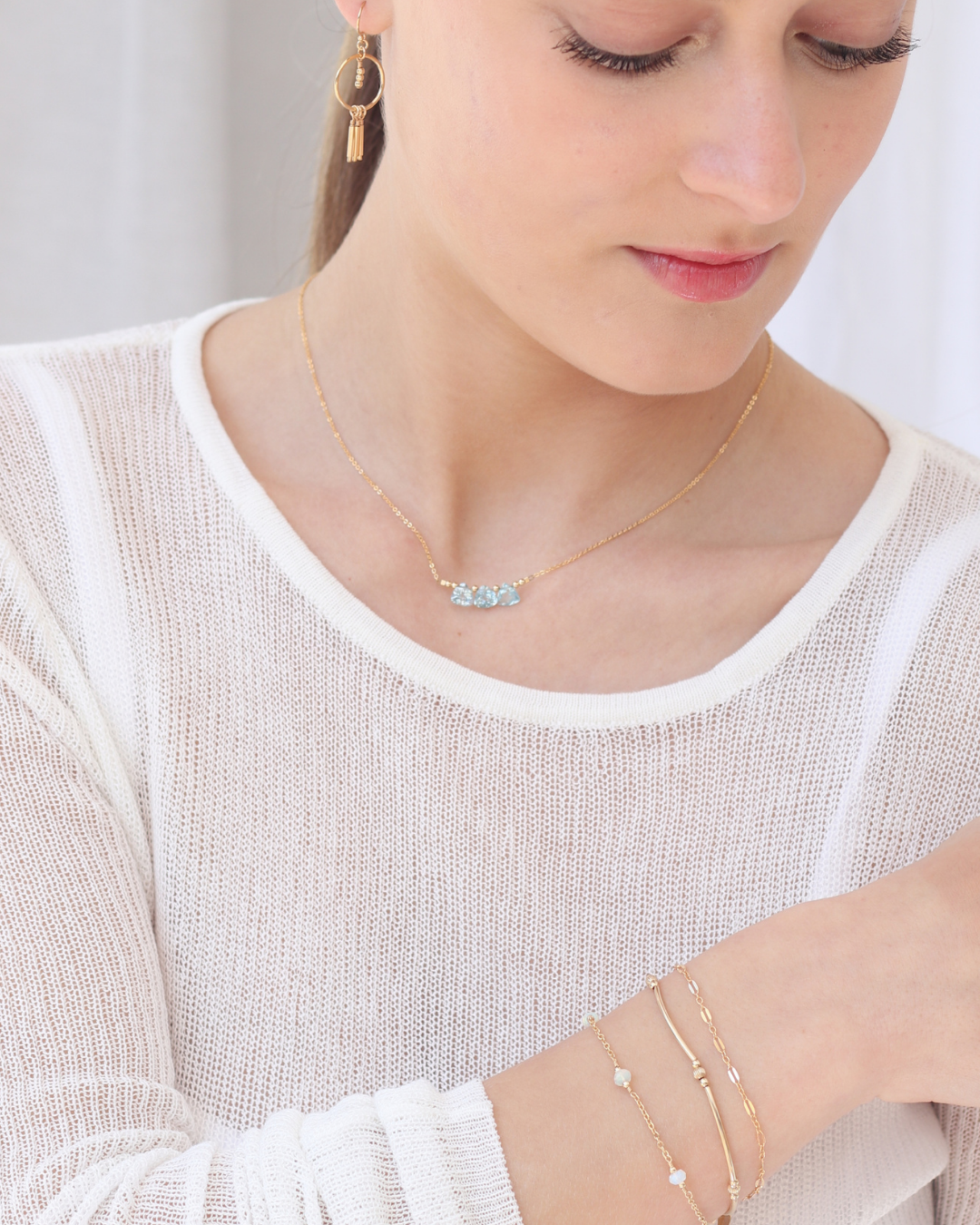 Woman wearing a white top featuring a gold necklace with blue topaz briolette gemstones, layered bracelets, and a pair of earrings.