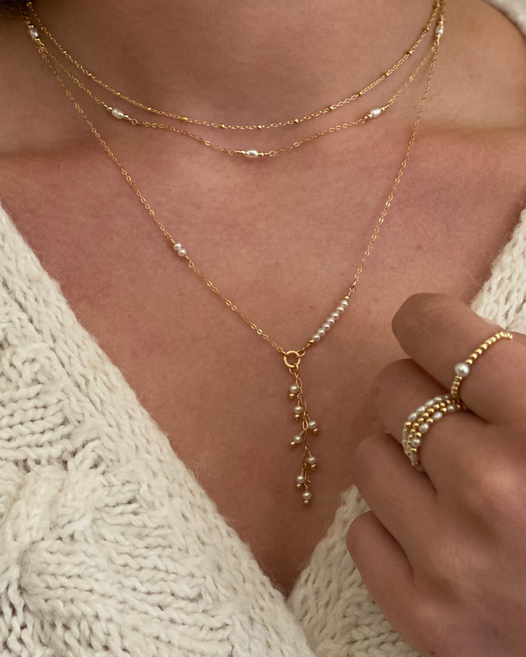 Close-up of a person wearing a gold necklace and ring on a beige sweater.