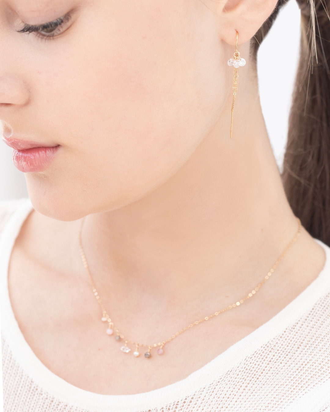 Close-up of a person wearing a delicate gold necklace with multiple gemstones and cubic zirconia thread earrings on a plain background