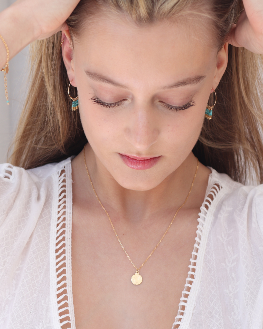 Woman wearing gold hoop earrings with turquoise gemstones and a gold pendant necklace on a neutral background