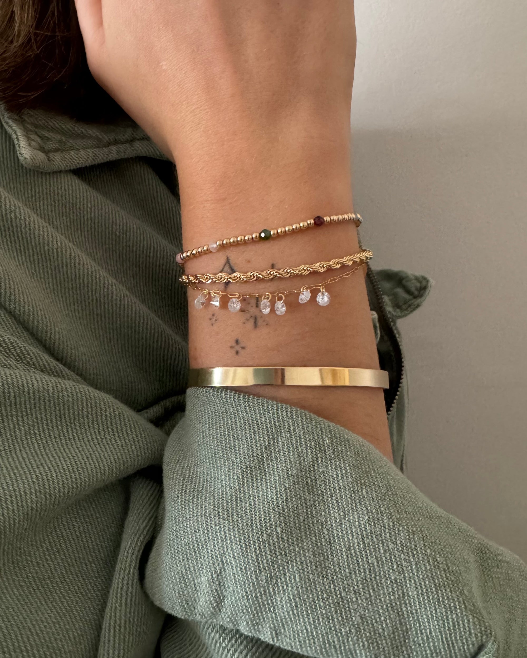Multiple gold bracelets, gemstones, cubic zirconias, bangle and rope chain bracelet on a wrist with a neutral background