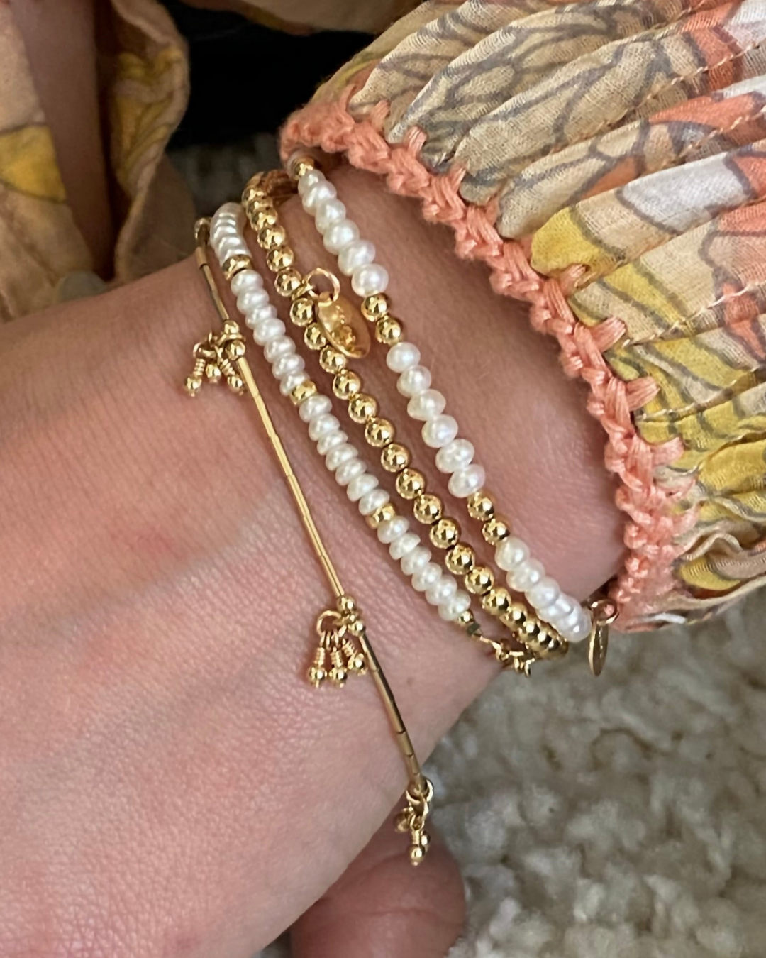 Close-up of a hand wearing multiple pearl and gold bracelets on a textured surface.