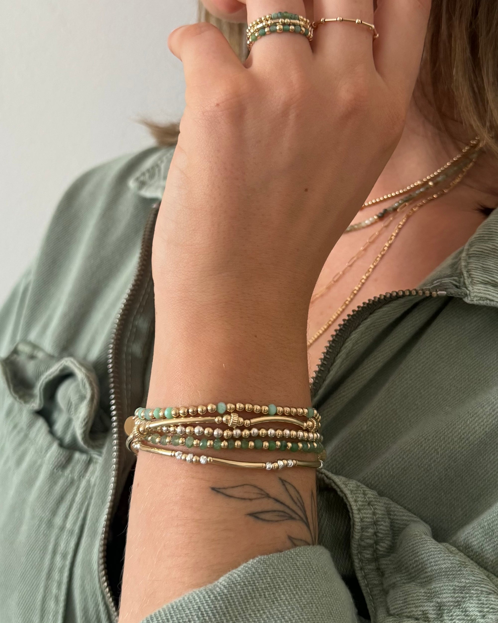 Close-up of a hand wearing multiple gold and  green gemstone bracelets and rings against a neutral background