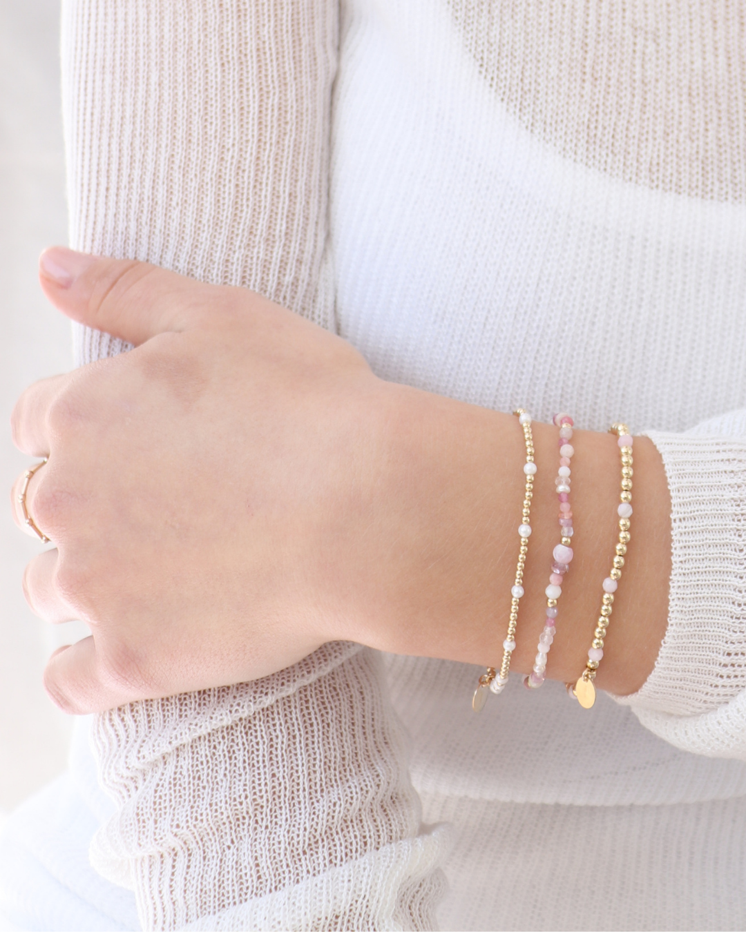 Close up of models hand wearing multiple gemstone beaded bracelets with pink peruvian opal gemstones on a neutral background