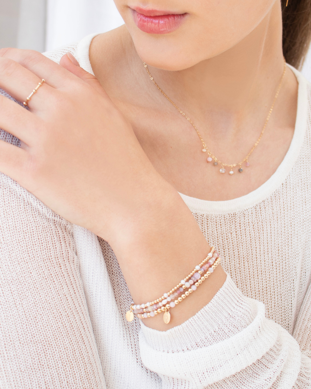 Close-up of a person wearing a stack of pink peruvian opal gemstone bracelets with a delicate gemstone necklace, and ring with a neutral background 
