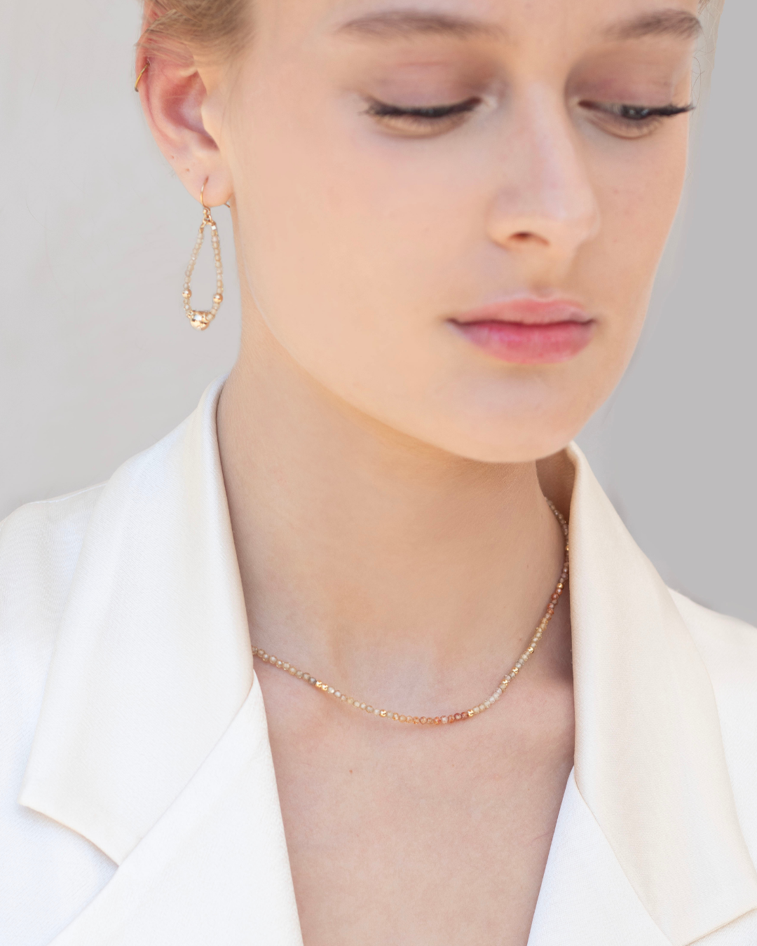 Close-up of a bride wearing a delicate gold and zircon gemstone necklace and gold and zircon earrings against a neutral background