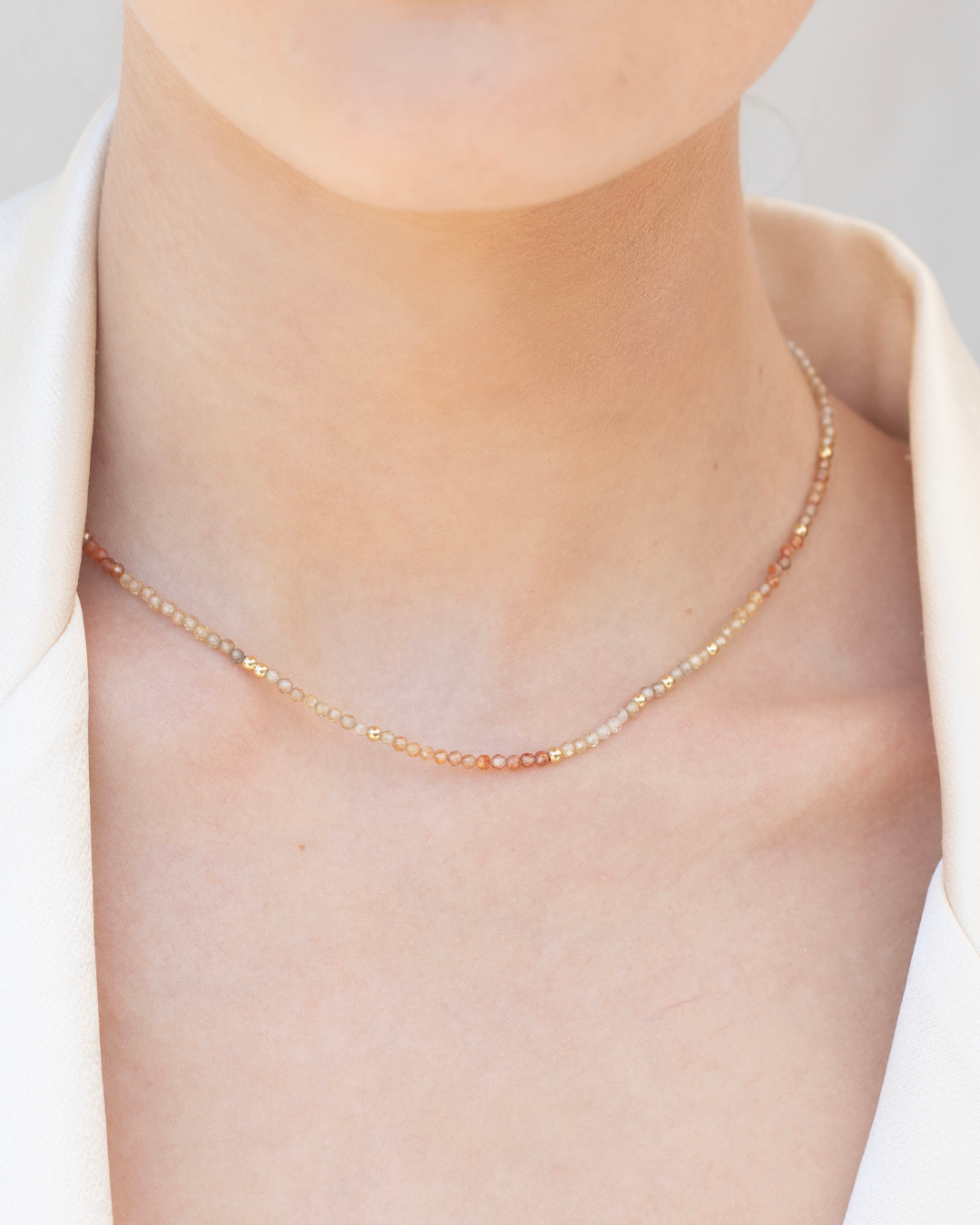 Close up of bride wearing a gold and zircon gemstone necklace with a cream satin blazer with a white background