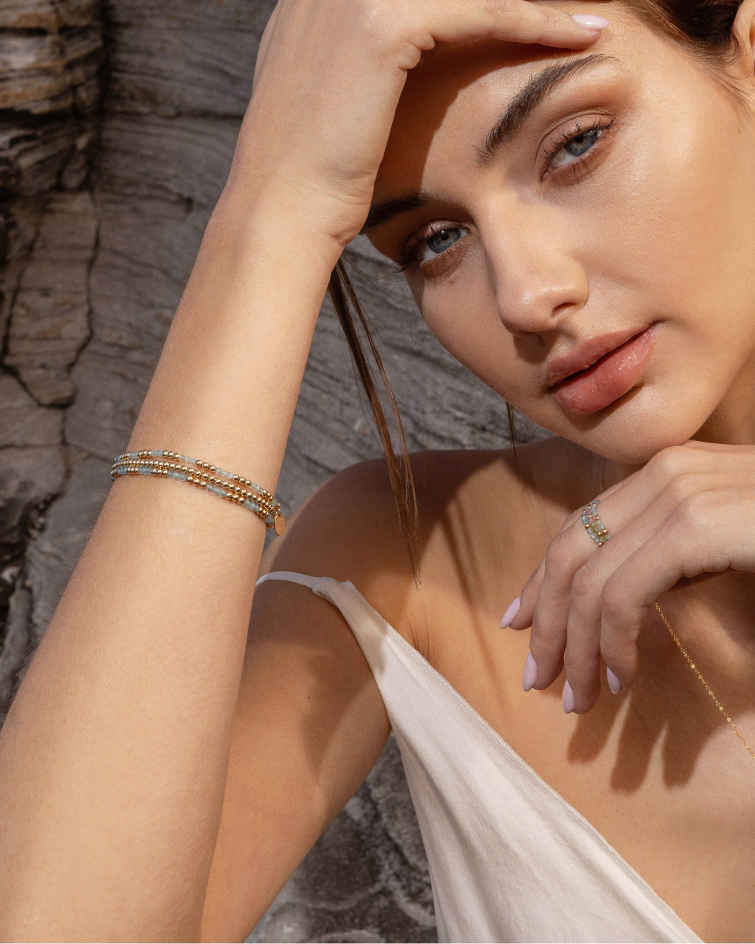 Woman wearing gold bead and gemstone bracelets with a stone wall background