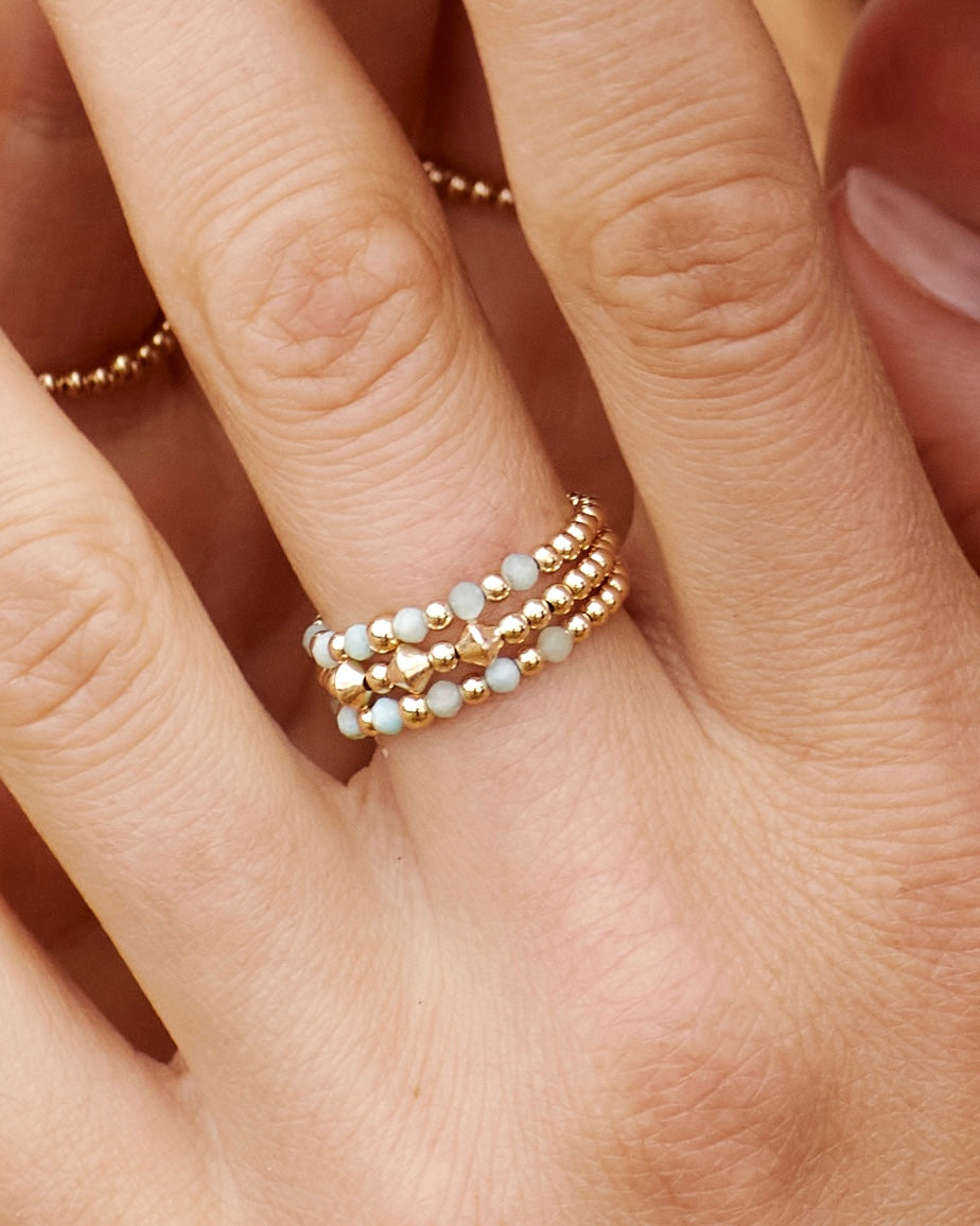 Close-up of a hand wearing multiple gold and larimar gemstone rings on a neutral background