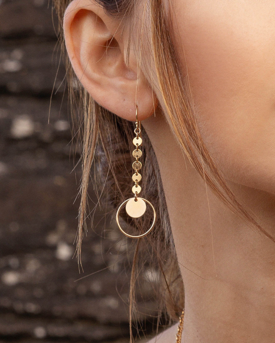 Gold earrings with circular pendants worn by a person against a blurred background