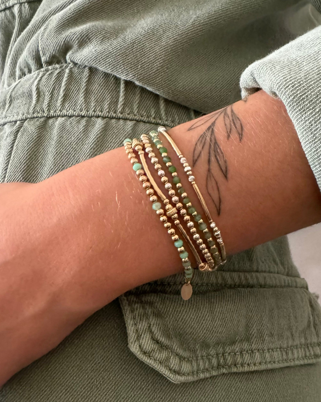 Close-up of a wrist with multiple bracelets and a tattoo on a person wearing a green jacket.