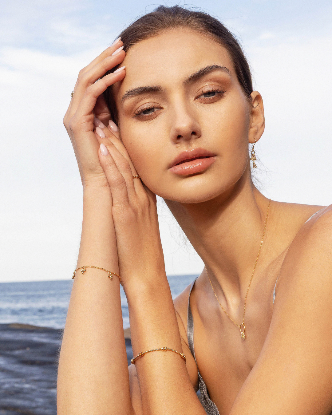 Woman on a beach with gold jewelry, wearing a grey silky strap top.
