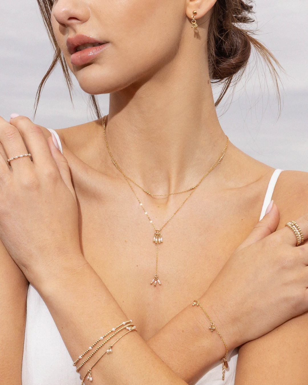 Woman wearing gold jewelry including a necklace, bracelet, and ring on a neutral background