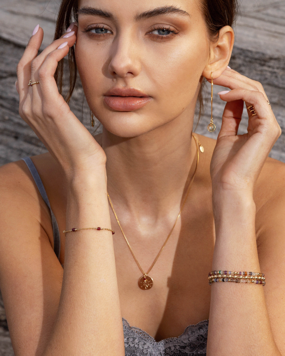 Woman wearing gold jewelry including a necklace, earrings, and bracelet against a neutral background