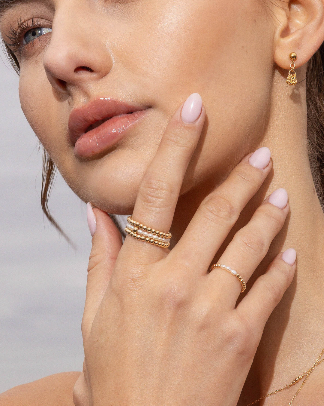 Close-up of a woman's face and hand wearing gold beaded rings on a neutral background