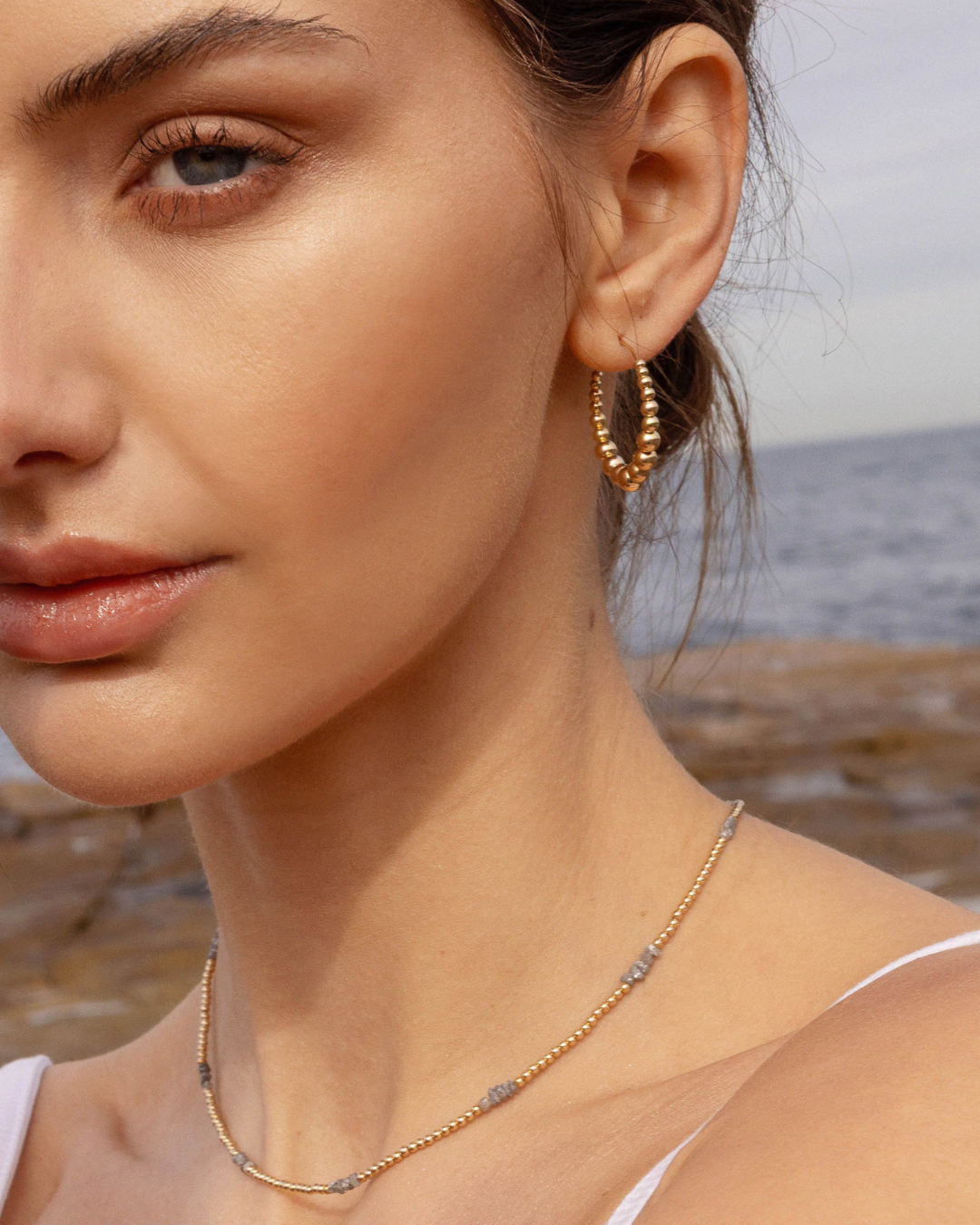 Close-up of a woman wearing gold hoop earrings and a necklace with a beach background