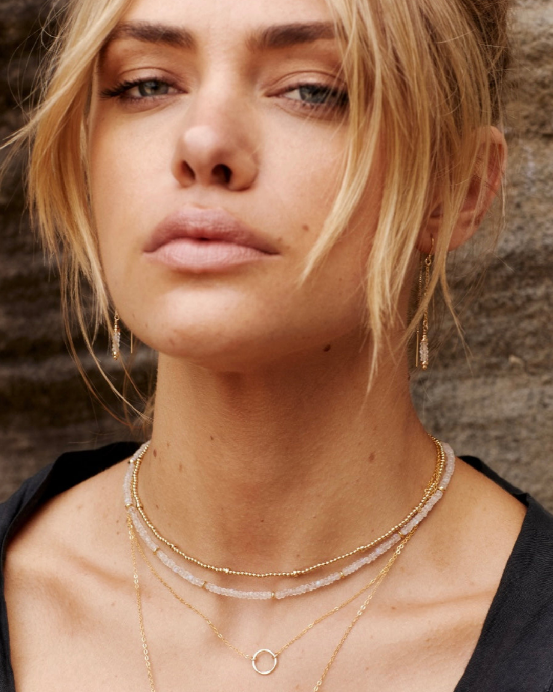 Close-up of a woman wearing gold necklaces against a stone wall background