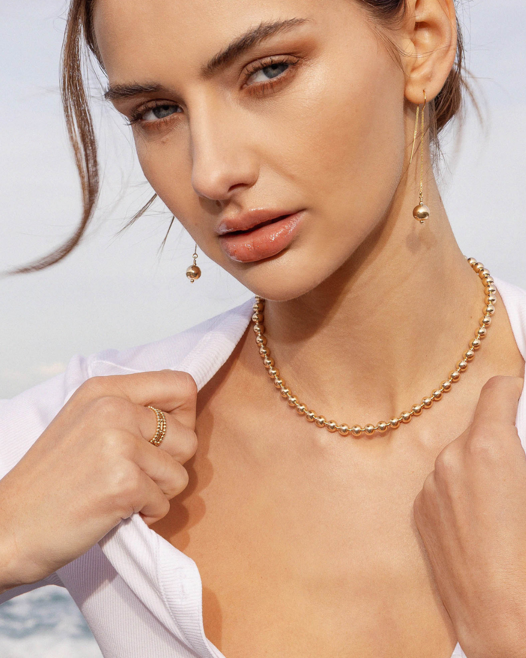 Woman wearing gold jewelry including a necklace, earrings, and ring against a light background