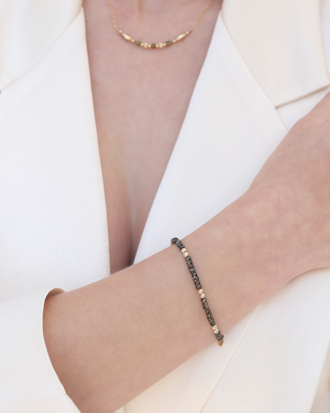 Close up of a woman wearing a gold and dark green zircon gemstone bracelet with and fine gold tourmaline gemstone necklace wearing a cream blazer.
