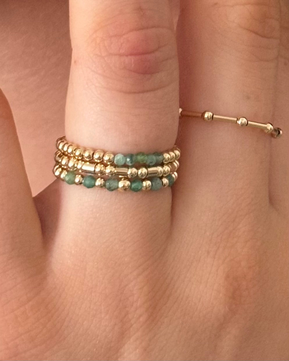 Gold and emerald beaded rings on a finger against a neutral background