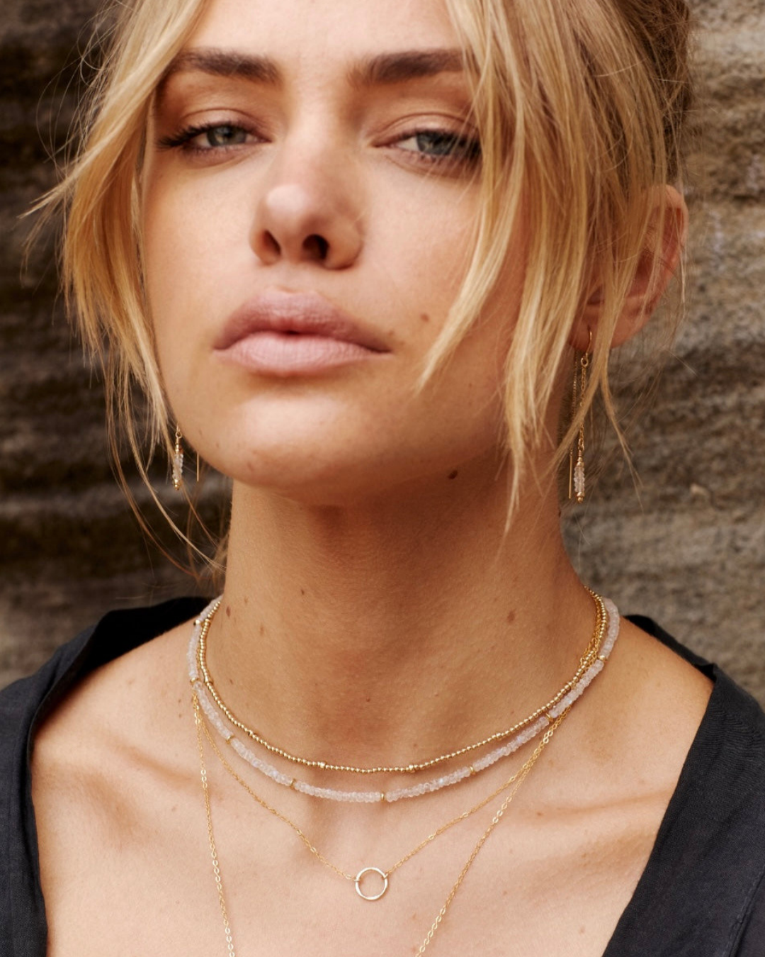 Close-up of a woman wearing gold and moonstone gemstone thread earrings and necklaces against a textured wall.