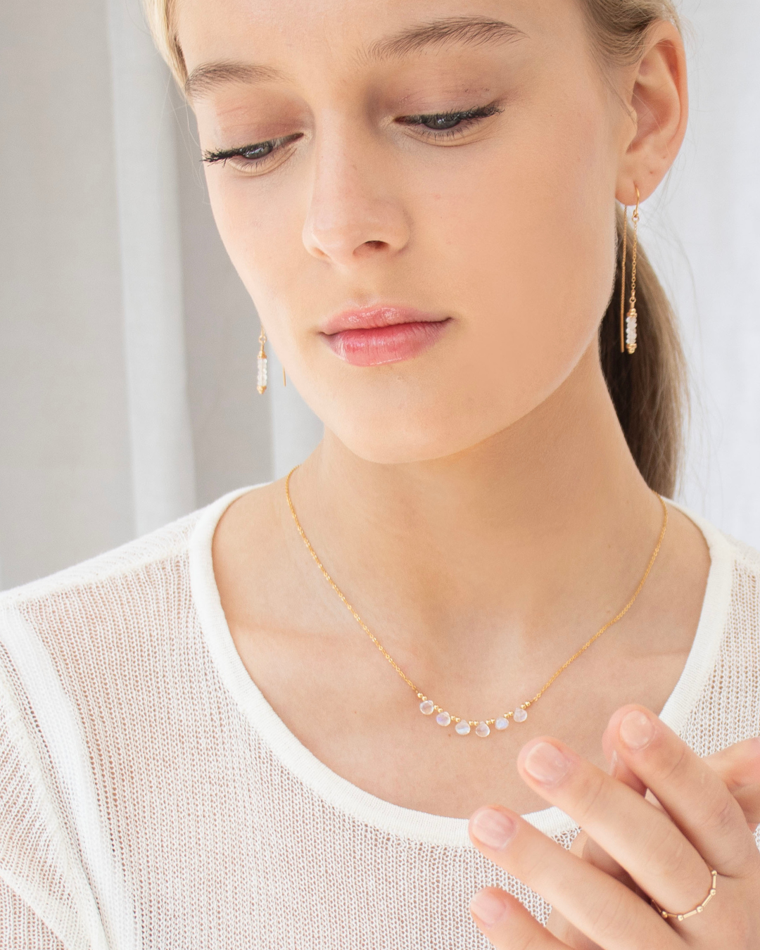 Close-up of a person wearing gold and moonstone gemstone thread earrings and  a delicate gold necklace with moonstone briolette gemstones on a neutral background. close-up of her face and upper body.