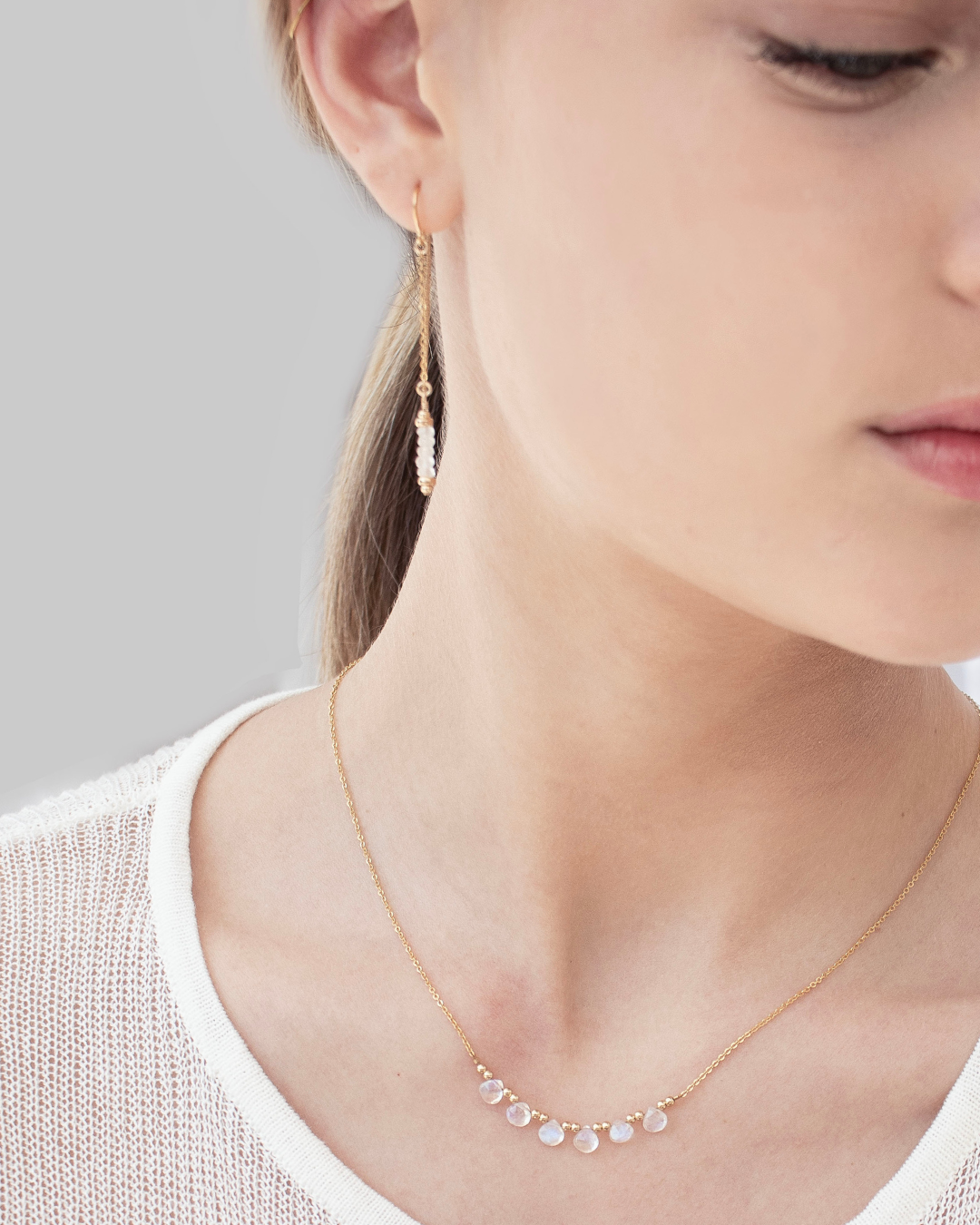 Close-up of a person wearing gold and moonstone gemstone thread earrings and  a delicate gold necklace with moonstone briolette gemstones on a neutral background.