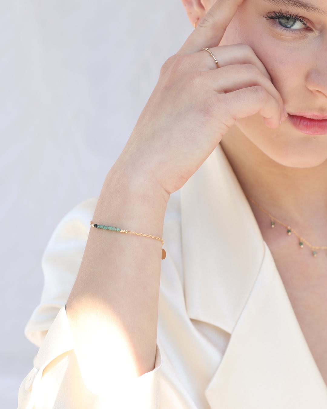 Woman wearing gold jewellery including a ring, emerald bracelet, and delicate emerald necklace on a light background