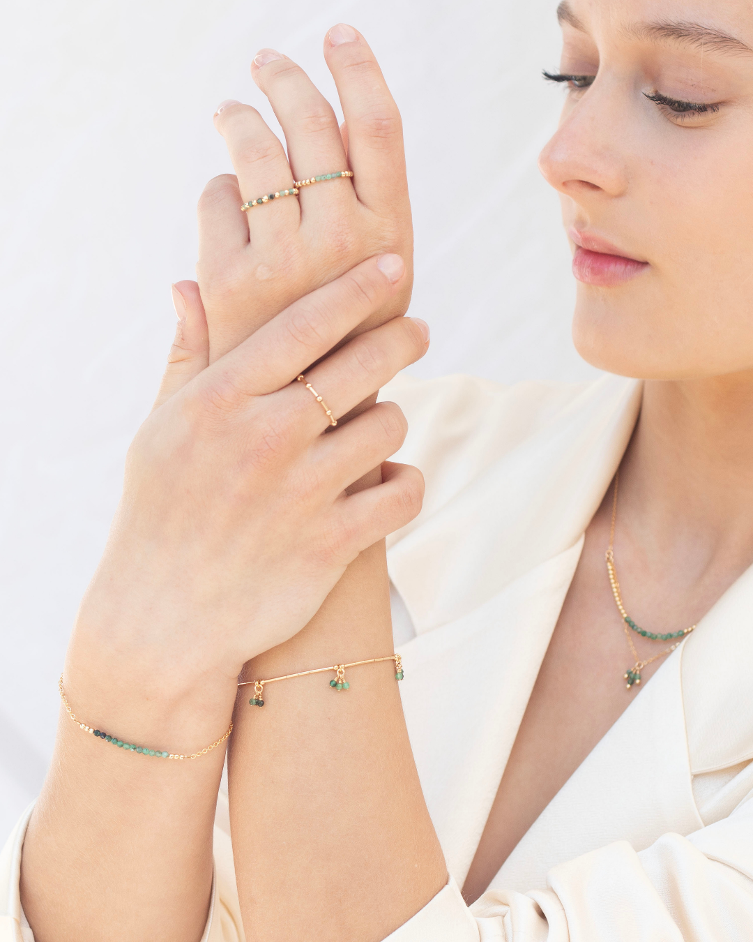 Woman wearing gold emerald jewellery including rings, bracelets, and a delicate necklace on a light background