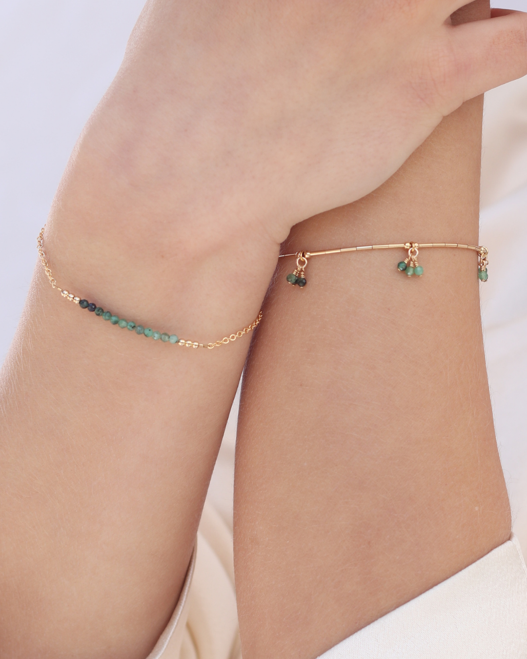 Close up of wrists wearing gold and emerald bracelets against a light background