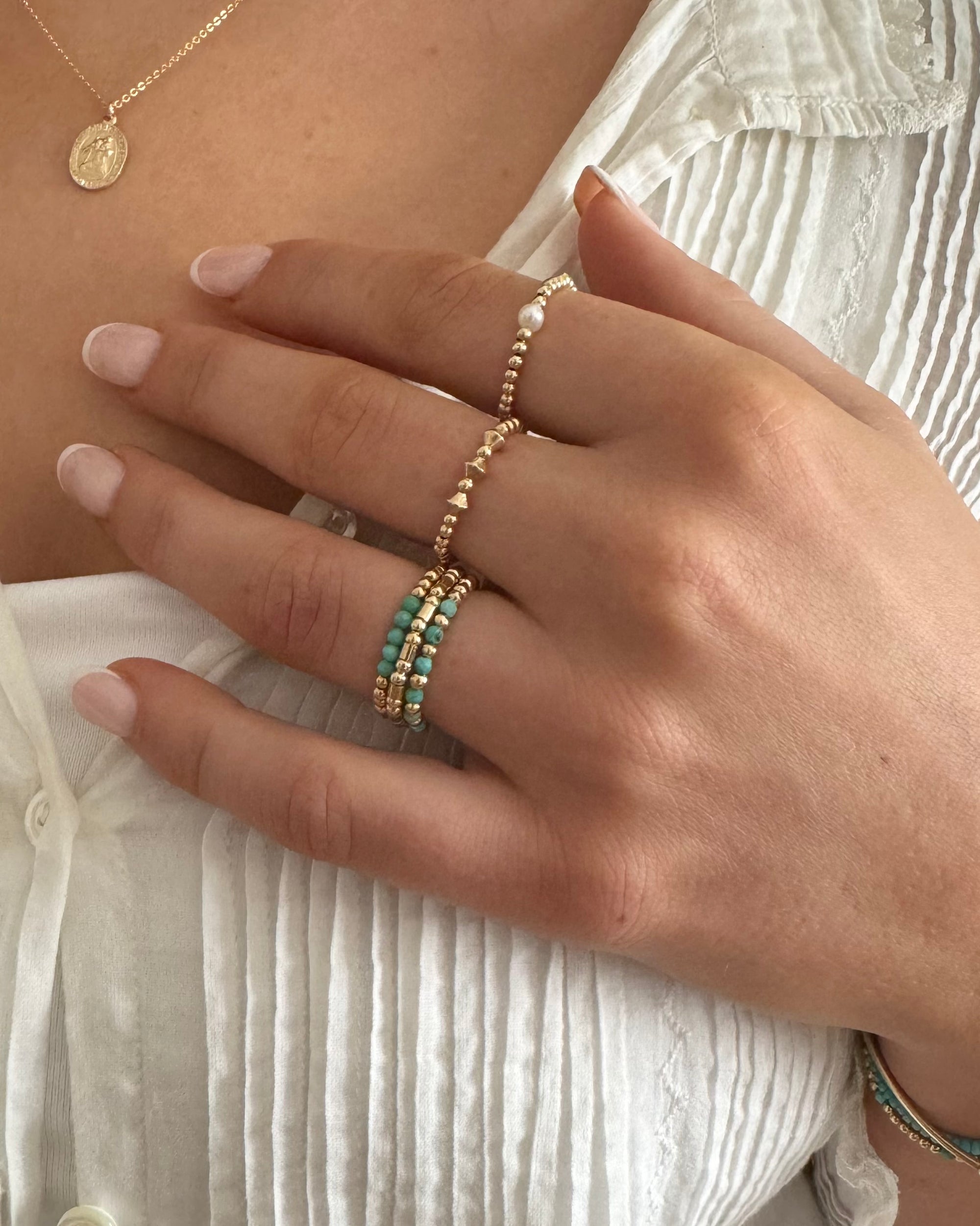 Close-up of a hand wearing gold rings with turquoise gemstones and pearl against a woman wearing a white blouse
