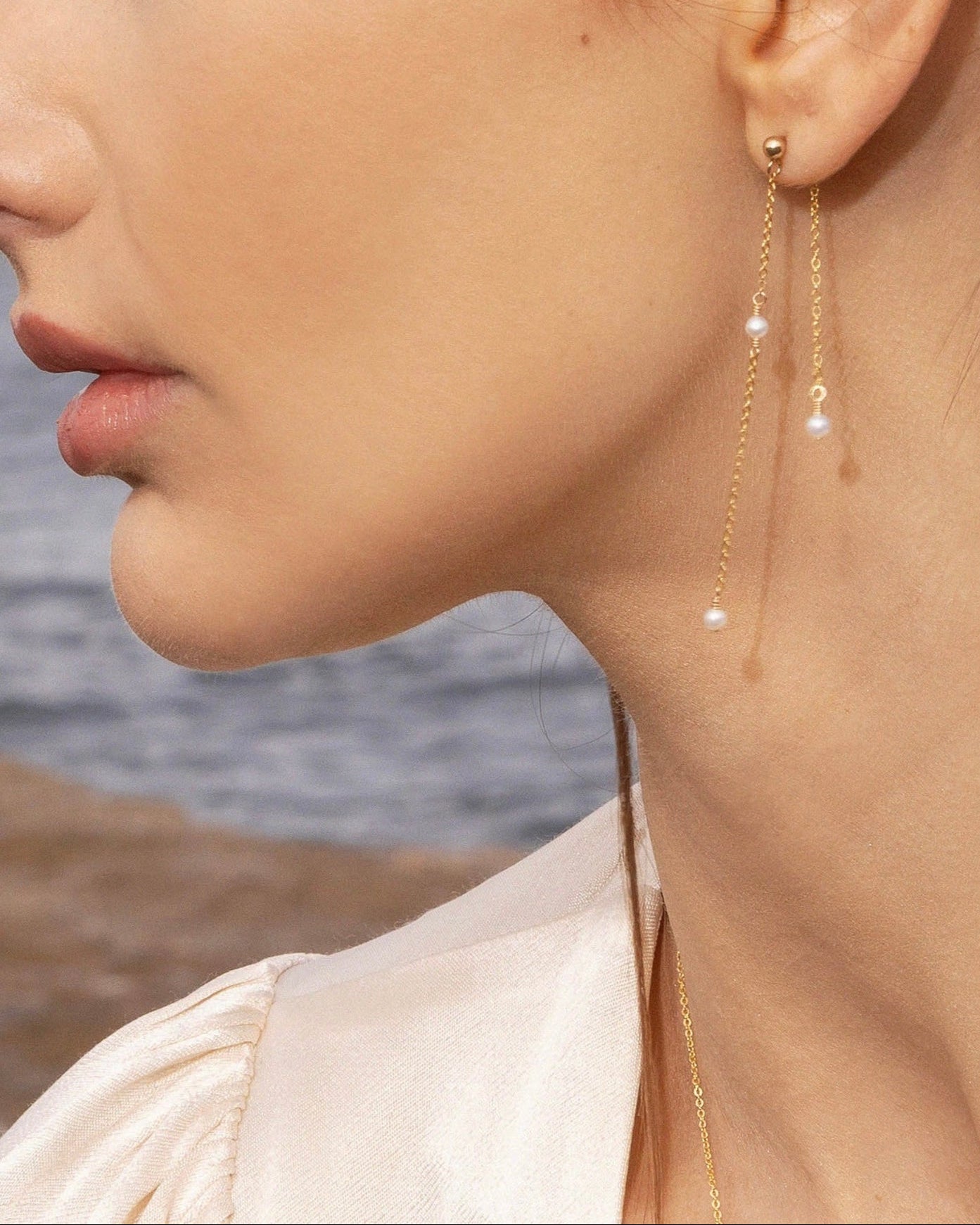 Close-up of a woman wearing gold earrings with pearl-like beads, set against a blurred natural background.