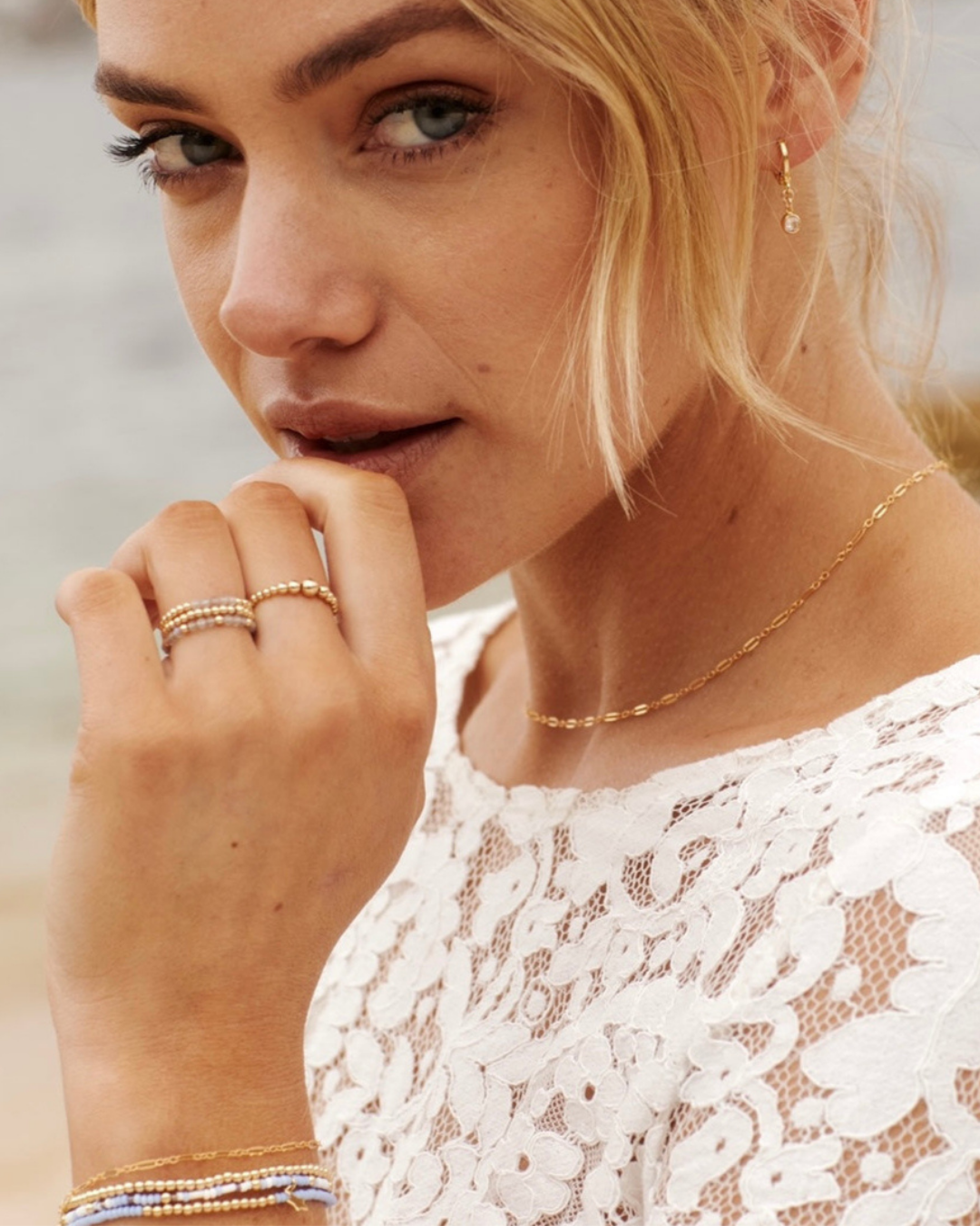 Woman wearing gold jewelry including a necklace, rings, and bracelets against a neutral background.