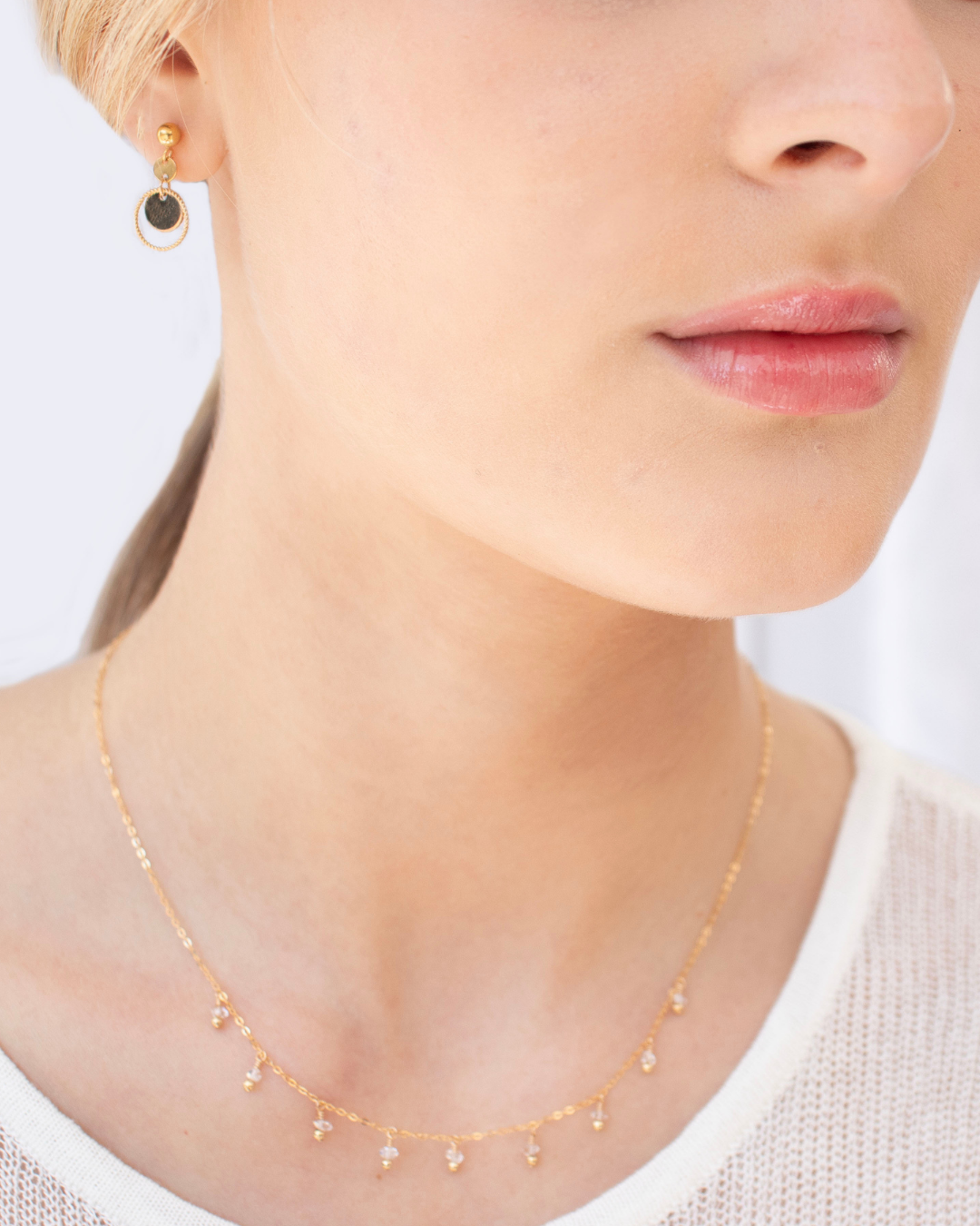 Close-up of a woman wearing a delicate gemstone gold necklace and gold disc earring wearing a white fine knit top