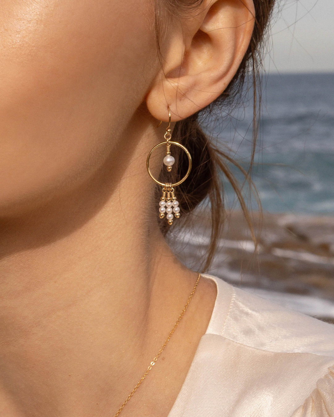 Close-up of a person wearing gold pearl drop earrings with an ocean background
