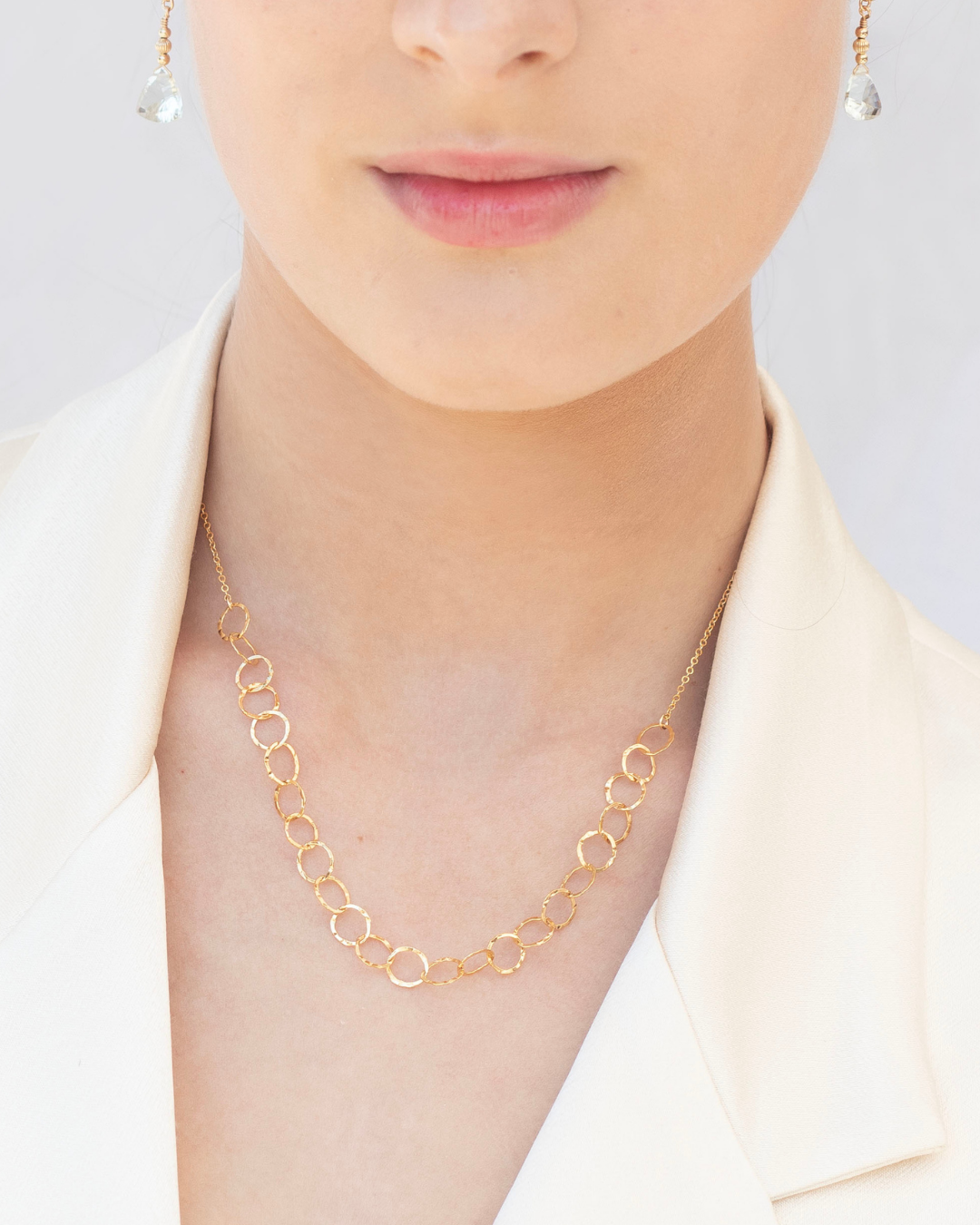 Gold necklace worn by a woman wearing a cream blazer with gold green amethyst earrings against a plain background