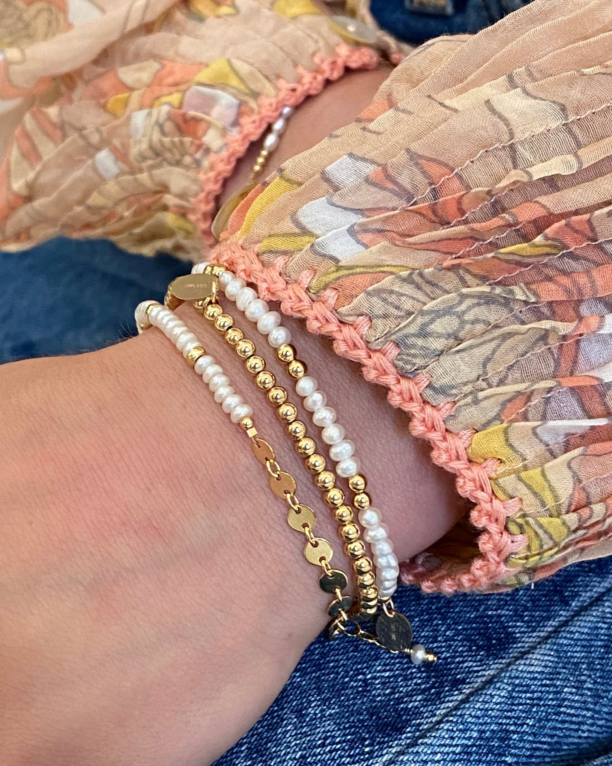 Close-up of a wrist wearing gold and pearl beaded bracelets with jeans and blouse sleeves in the background.