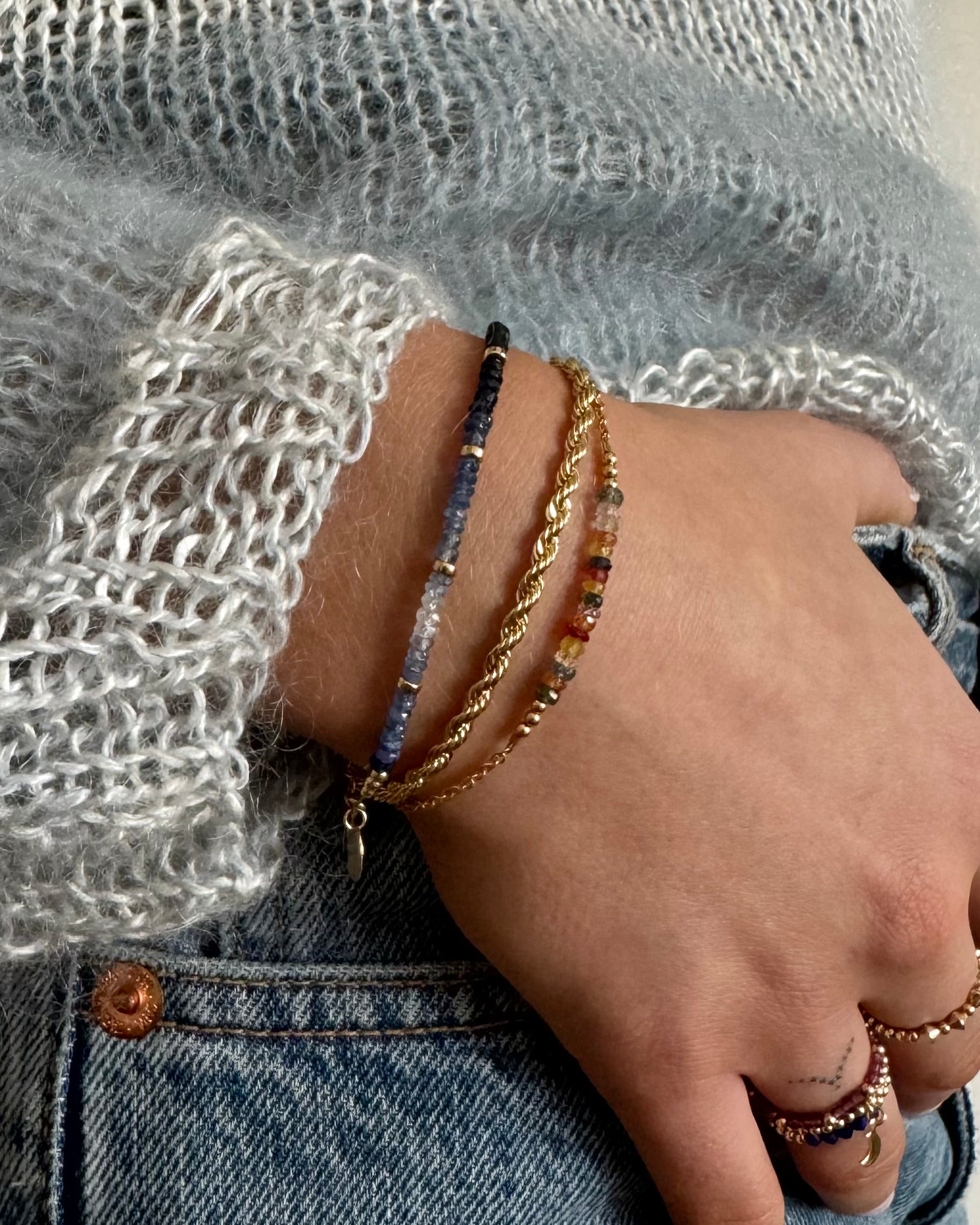 Hand wearing multiple gold sapphire bracelets against jeans and a jumper