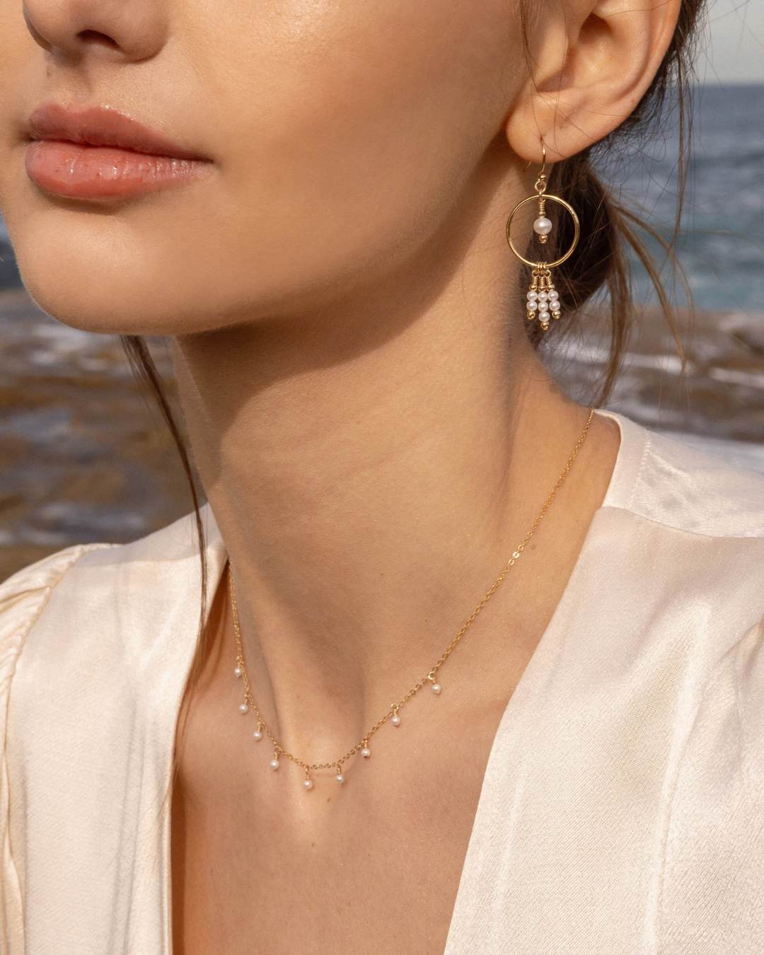Close-up of a woman wearing a delicate pearl gold necklace and earring with a blurred beach background