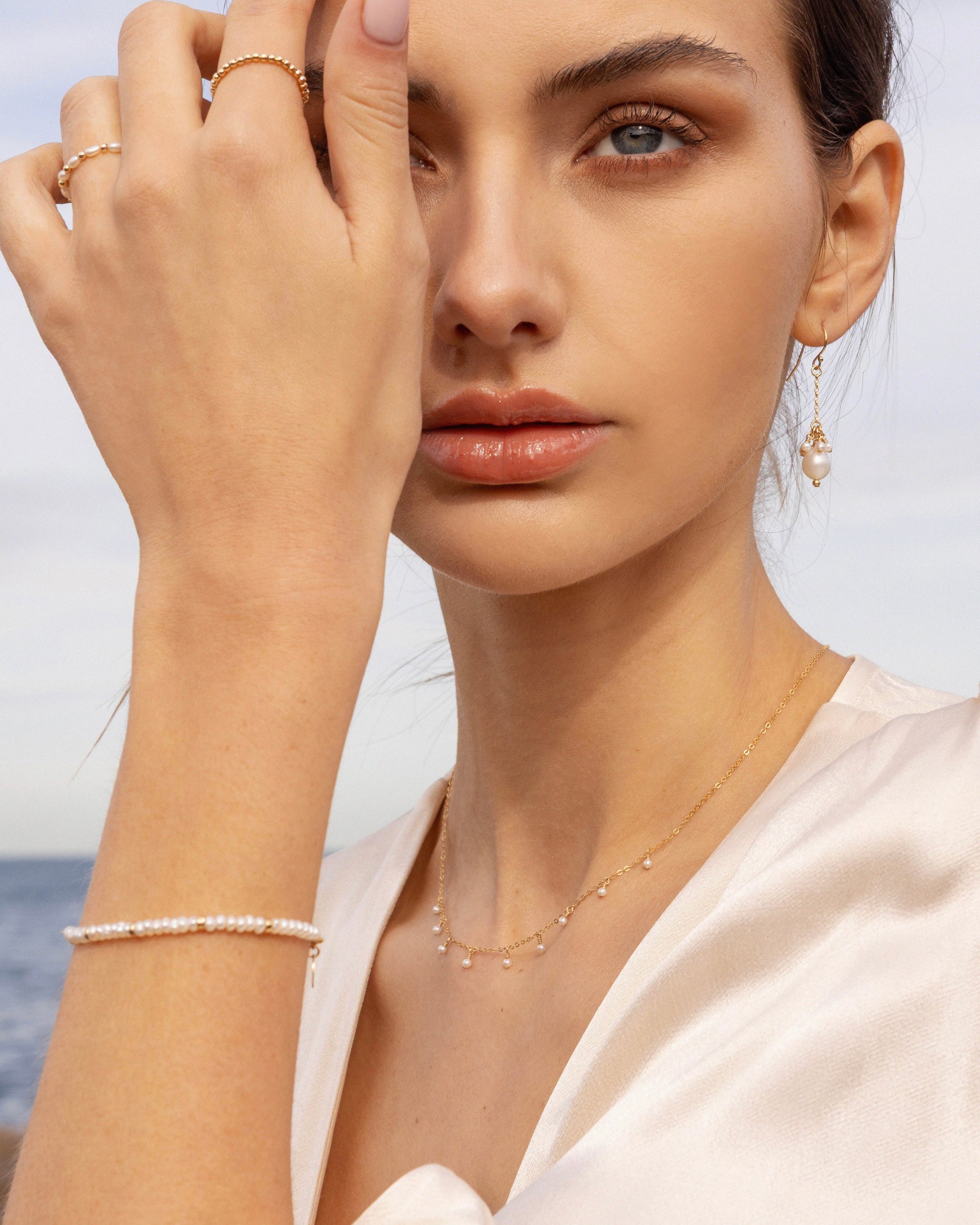 Woman wearing gold pearl jewellery, including necklace, earrings, bracelets and rings in a cream satin top with a blurred ocean background