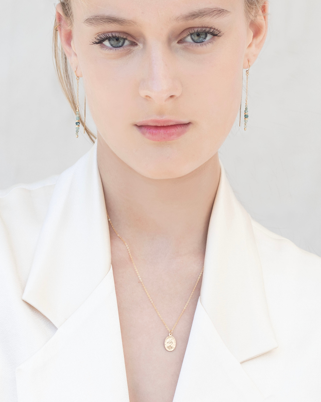 Woman wearing a gold necklace featuring a Saint Christopher pendant and grandidierite gemstone earrings and cream sating blazer