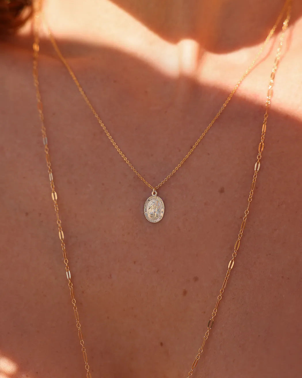 Close-up of a person wearing a gold necklace with a coin pendant featuring St Christopher, suspended from the fine cable chain