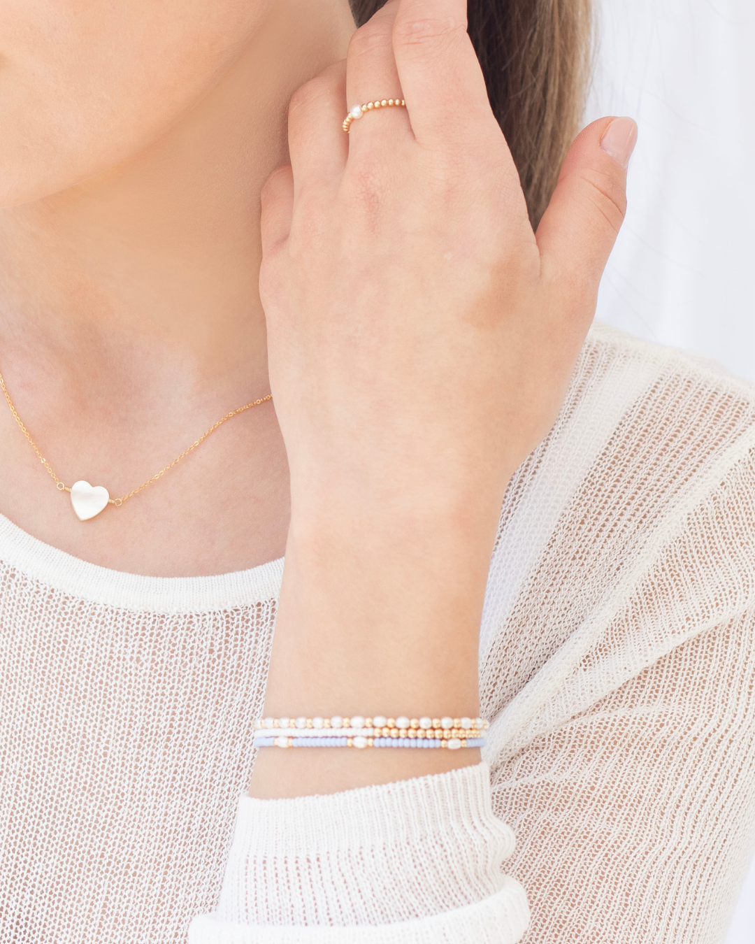 Close-up of a woman wearing gold and white beaded bracelets, pearl ring and a  mother of pearl heart pendant necklace and fine white knitted top.