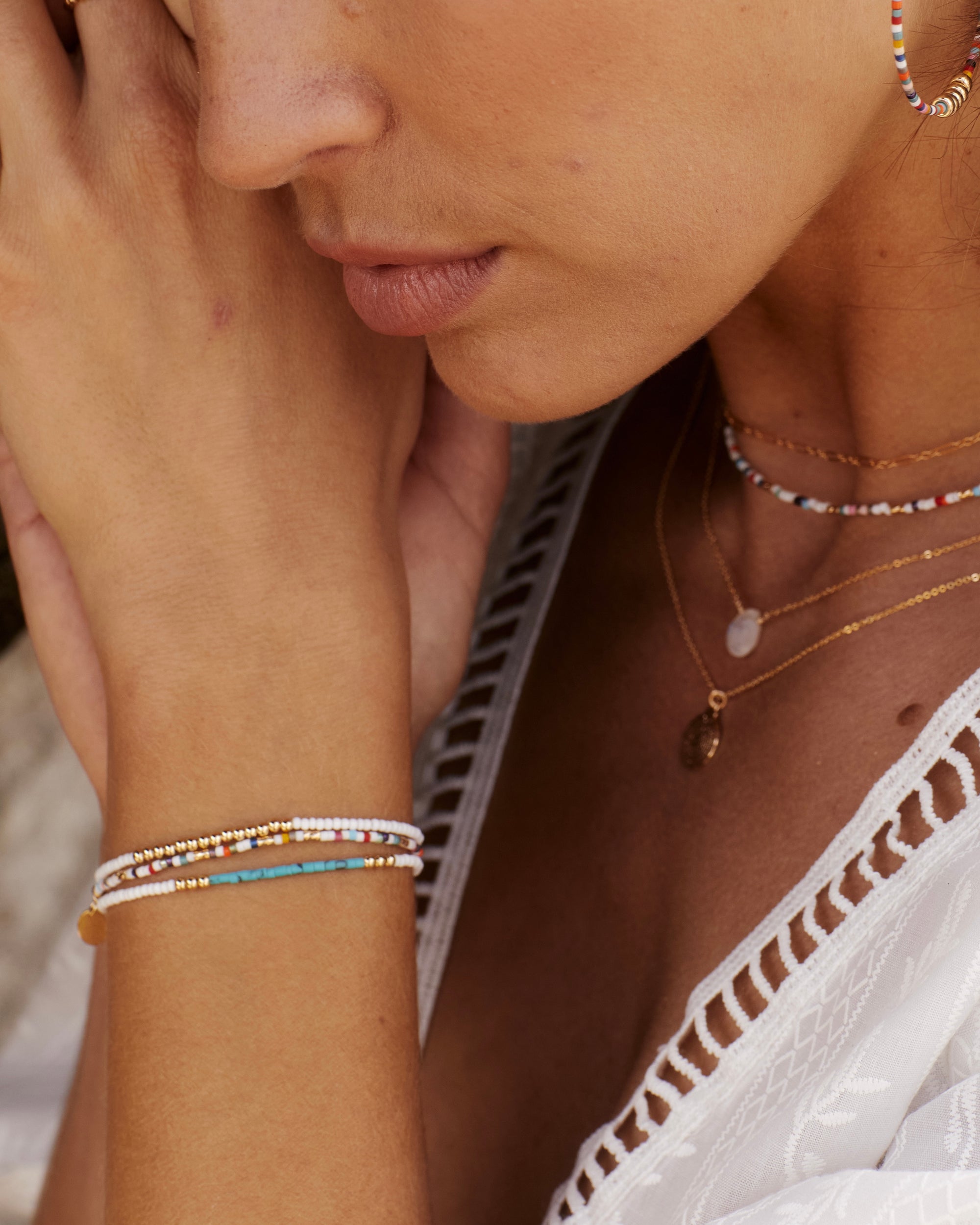 Woman wearing a stack of gold and white beaded bracelets, hoop earrings and layered necklaces.