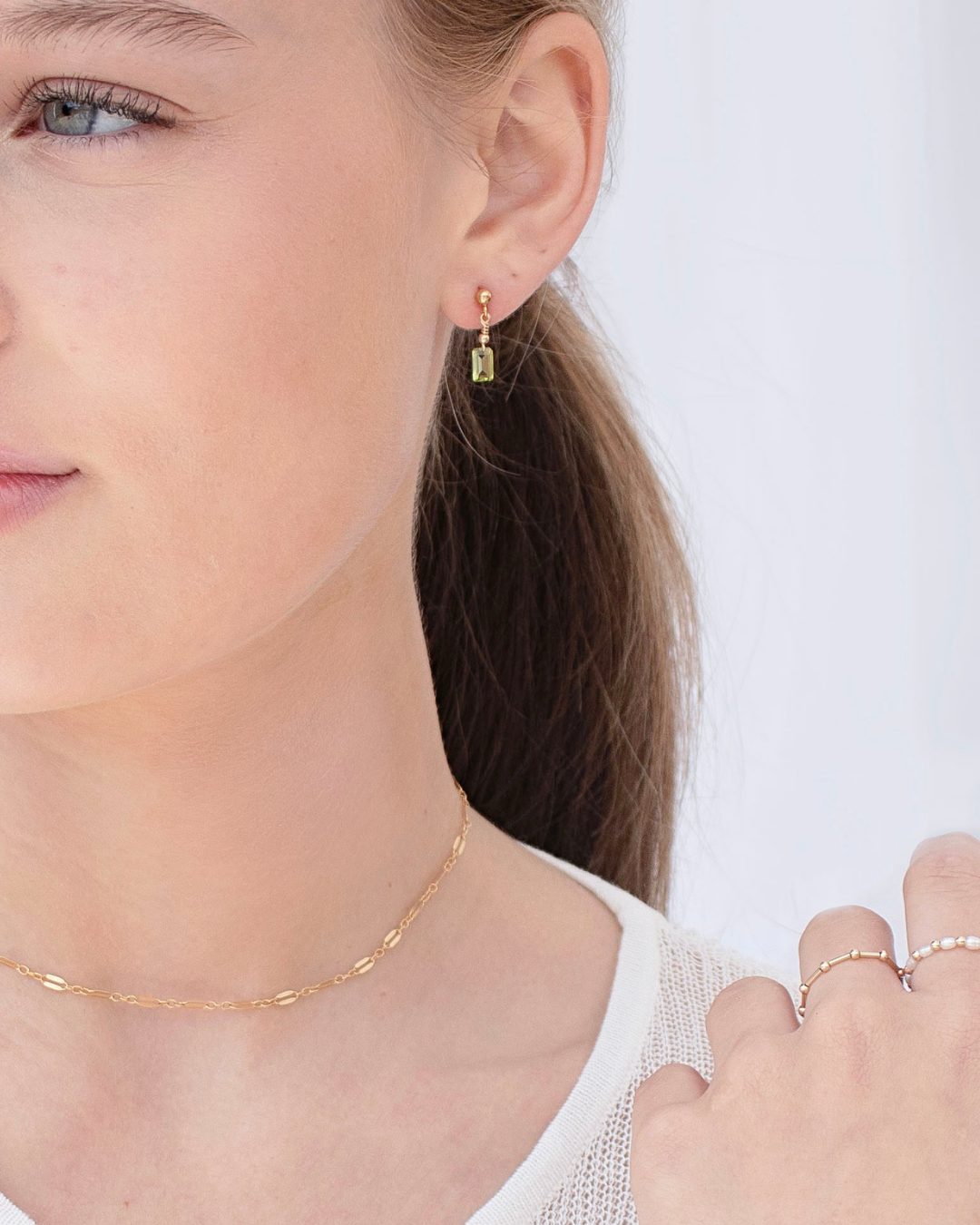Close-up of a woman wearing gold peridot earrings, gold delicate necklace and gold pearl beaded rings.