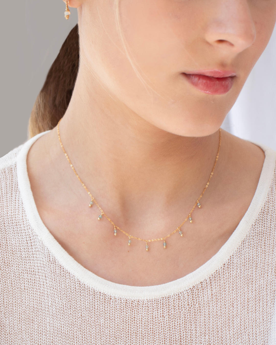 Close-up of a woman in a fine white knit top wearing a fine gold necklace with apatite gemstones 