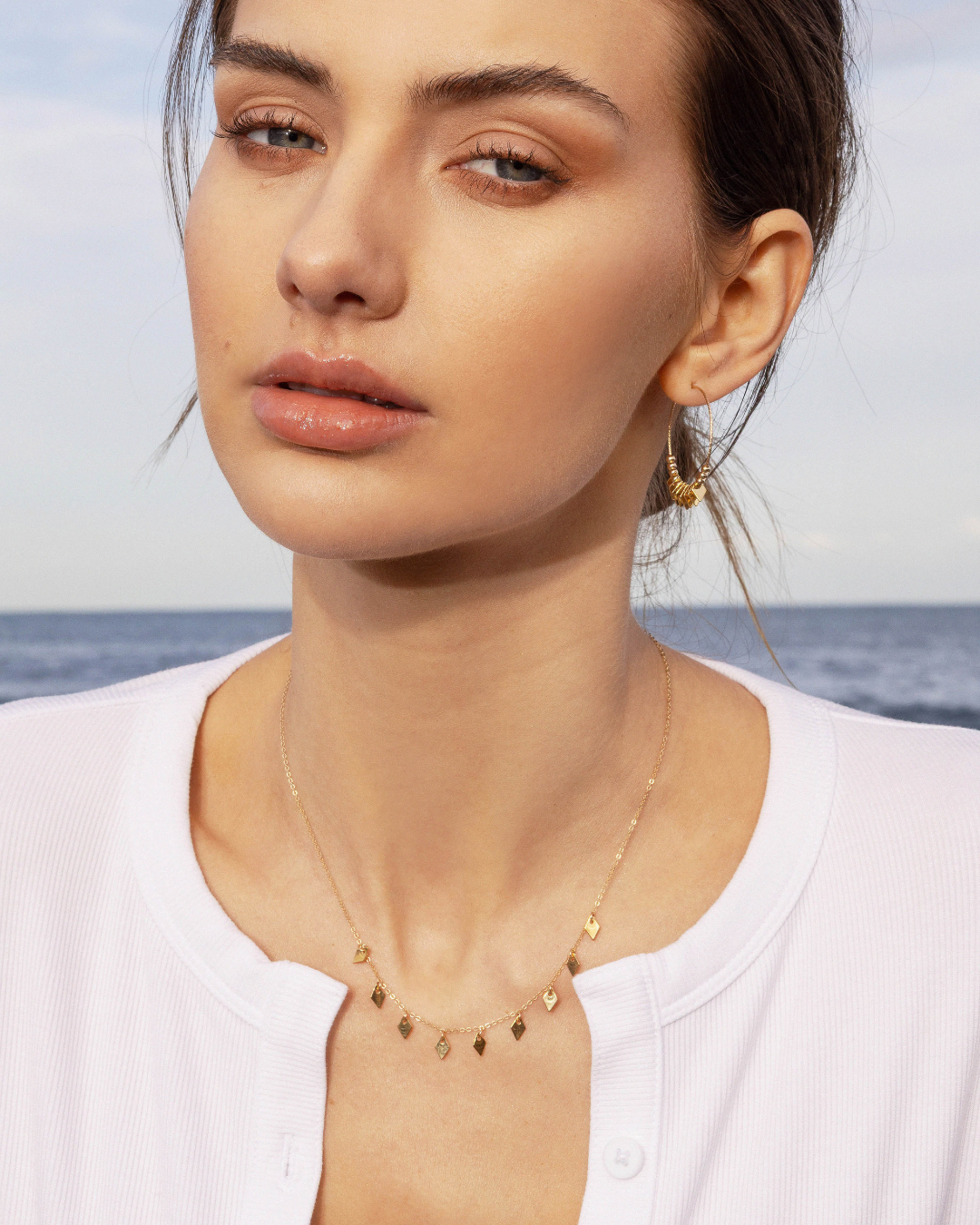 Woman wearing a gold necklace with diamond charms and matching hoop earrings with a blurred ocean background