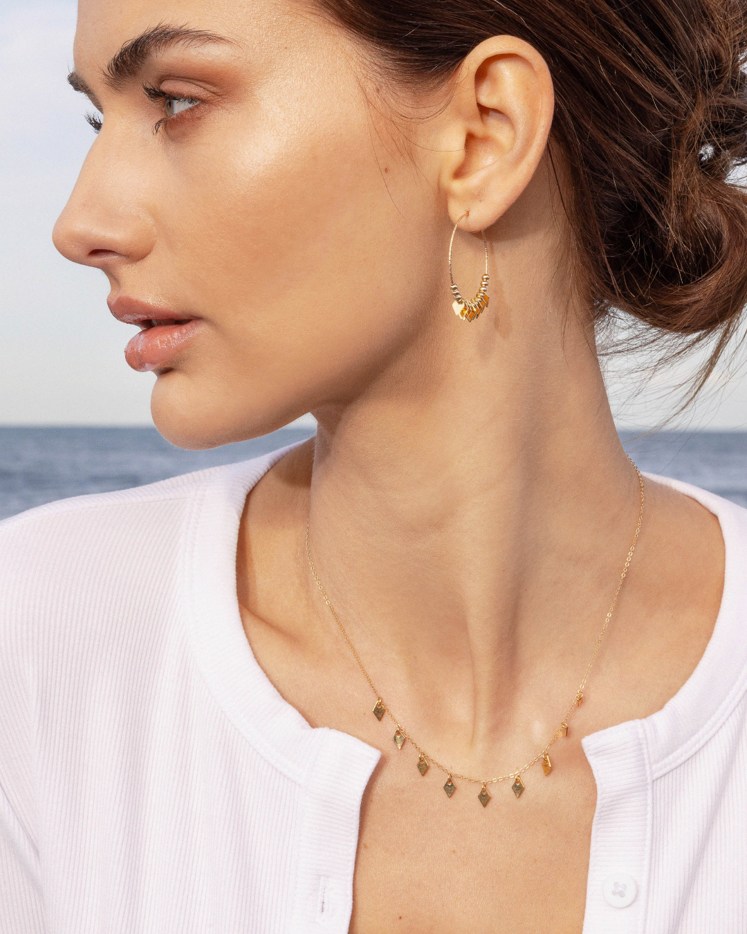 Close up of a woman wearing gold hoop earrings and a delicate necklace with a blurred ocean background