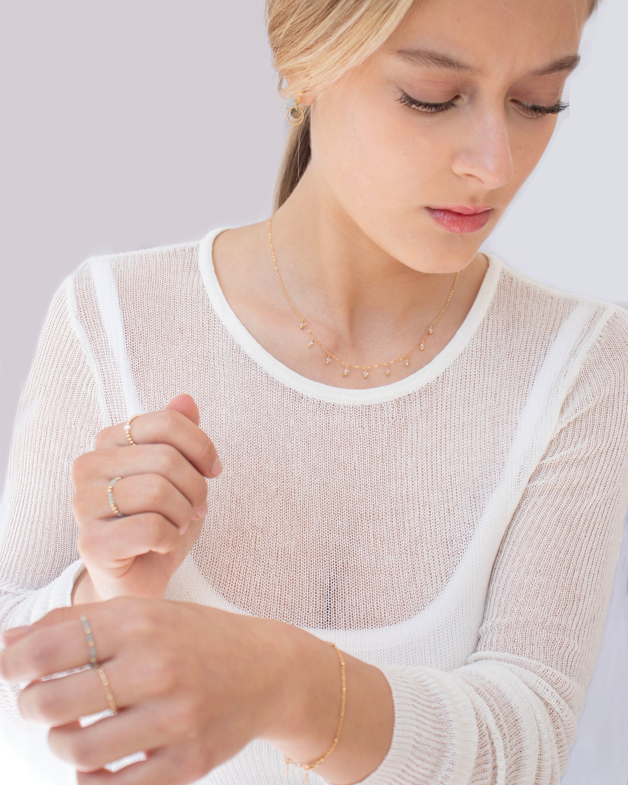 Woman wearing a sheer white top with gold jewellery including rings, bracelet, earrings and a herkimer diamond necklace,.